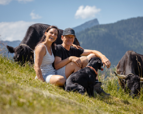 Lächelndes Paar mit Hund auf einer Almwiese, umgeben von schwarzen Kühen, mit Berglandschaft im Hintergrund.
