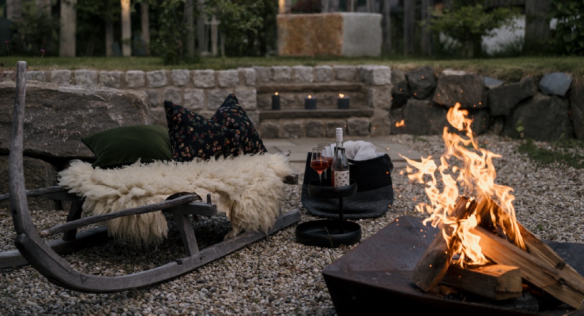 Gemütlicher Sitzbereich im Garten mit Holzschlitten, Feuerstelle, Kerzen und Getränken bei Abendstimmung.