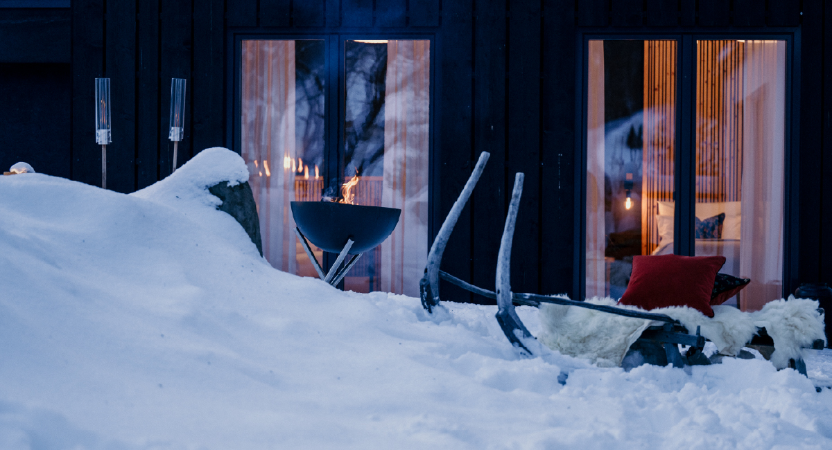 Verschneite Terrasse mit Feuerstelle, traditionellem Holzschlitten und rotem Kissen vor einer gemütlich beleuchteten Hütte.