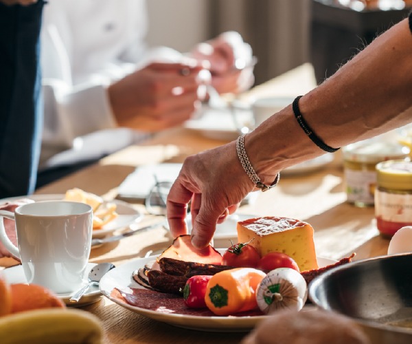 Reich gedeckter Frühstückstisch mit Käse, Wurst, frischem Gemüse und einer Hand, die zu einer Scheibe greift.